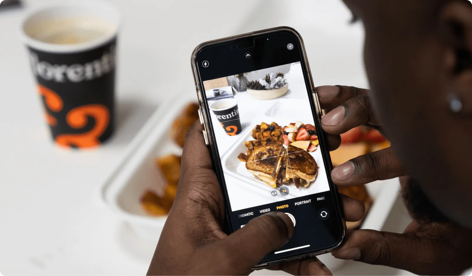 Person holding a smartphone, taking a photo of a meal with French toast, potatoes, and fruit, with a coffee cup in the background.