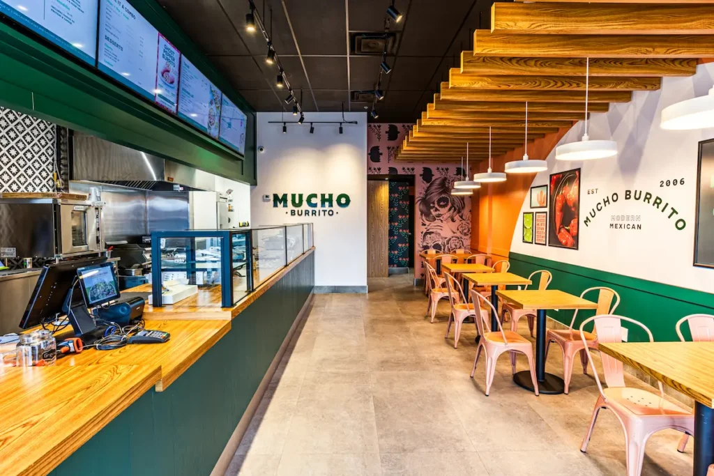 Modern Mucho Burrito interior with wooden ceiling accents, Catrina mural, colourful metal seating, and open kitchen counter.