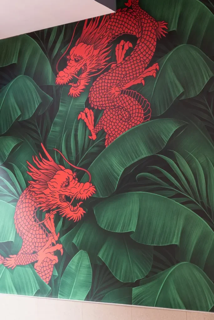 Decorative wall mural inside a Manchu WOK restaurant featuring two red dragons set against a backdrop of large tropical green leaves.