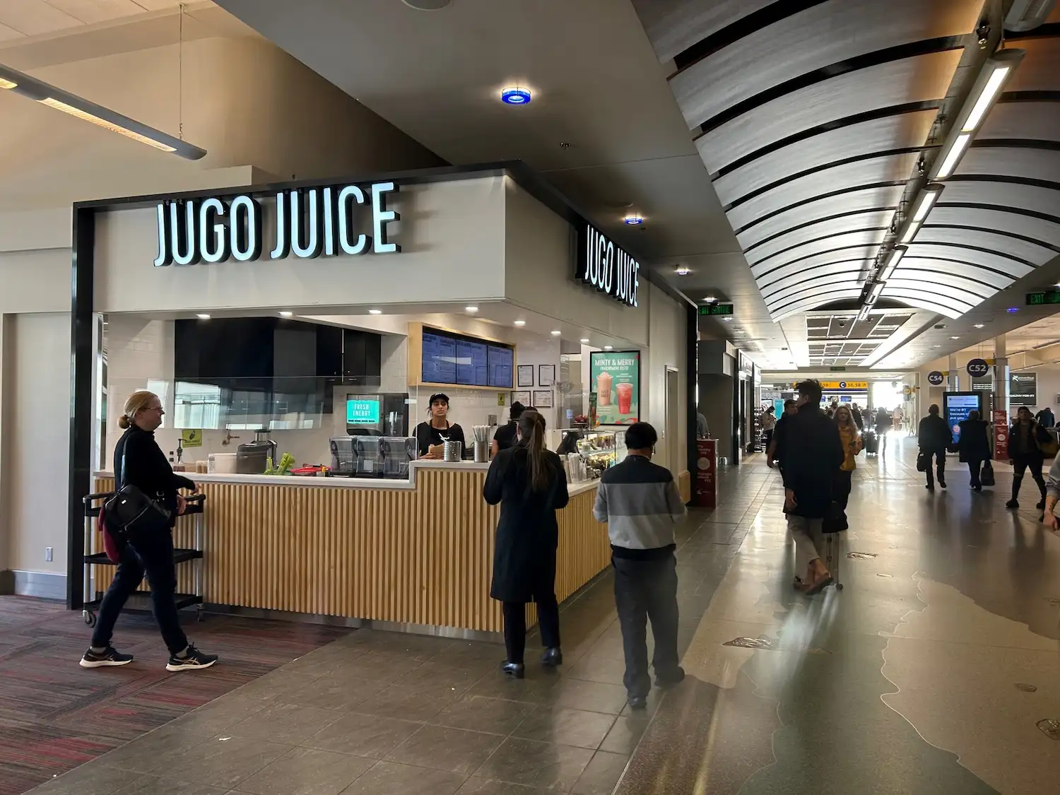 A Jugo Juice kiosk in the concourse of Calgary International Airport, with customers ordering at the counter and travellers walking past the gate areas in the background.