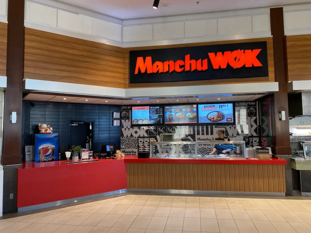 A Manchu WOK food court counter with a red service bar, digital menu boards in French, and a staff member preparing dishes behind the sneeze guard.