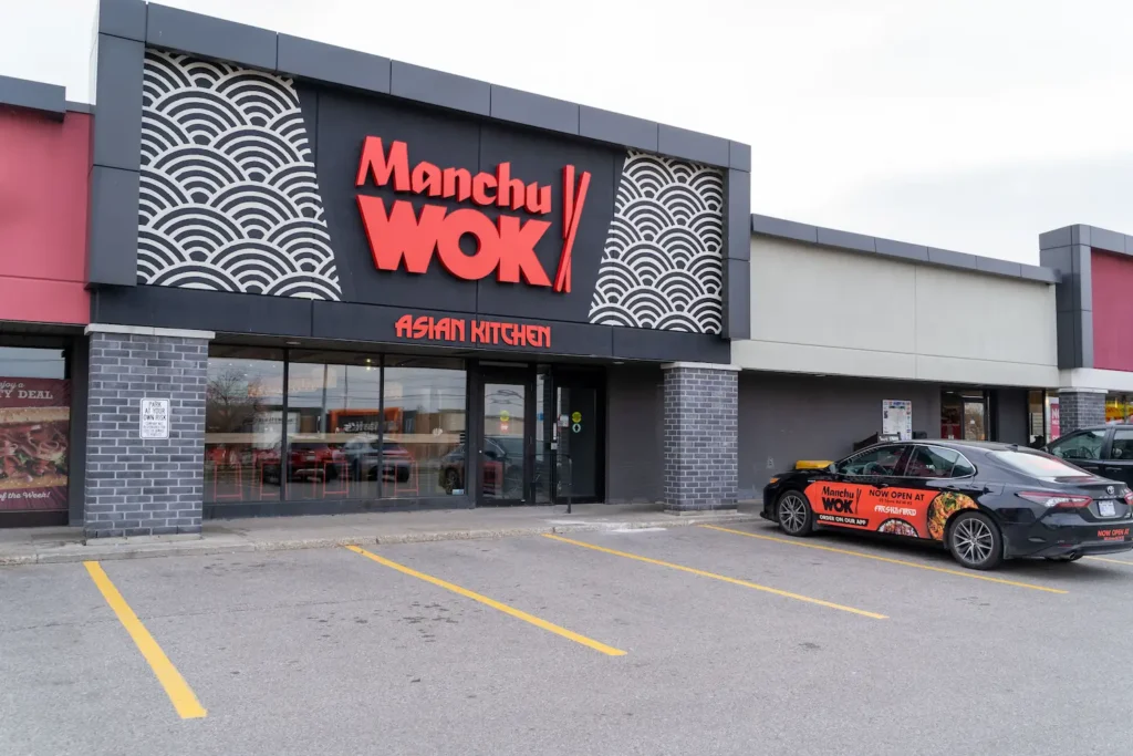 Exterior of a Manchu Wok Asian Kitchen restaurant in a strip plaza, with a large red logo sign on a dark facade and a branded delivery car parked out front.