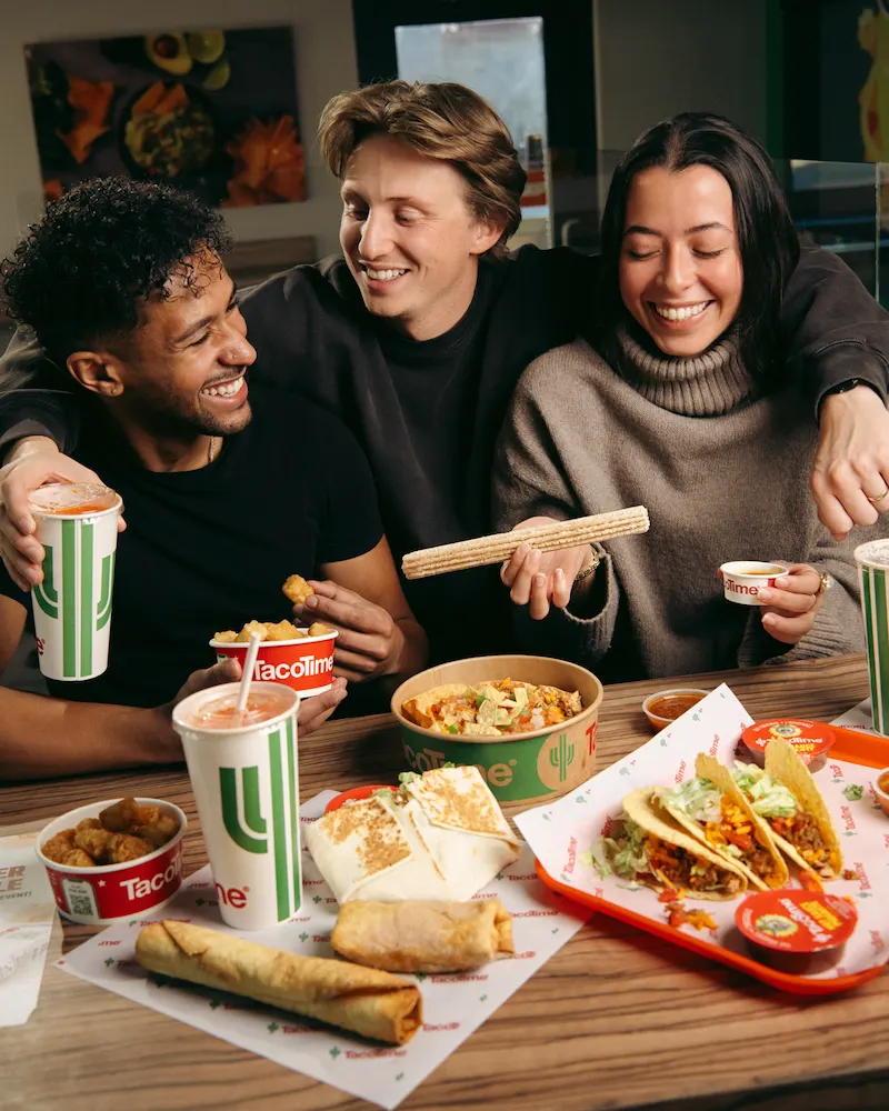 Three friends sharing tacos, burritos and Mexi-Fries at a table inside a TacoTime restaurant.